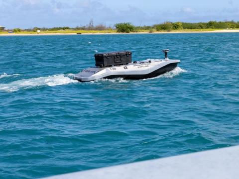  An unmanned surface vessel moves out to sea from Sand Island near Honolulu to deliver parts for a 3D printer aboard the Wasp-class amphibious assault ship USS Essex (LHD-2) during Exercise Rim of the Pacific 2026, July 7, 2026. Although there is no human operator aboard such a vessel, there is a human footprint—and often a very large one—needed to operate and maintain an “unmanned system,” the author says. Credit: U.S. Navy photo by Mass Communications Specialist 3rd Class Charles Harbour