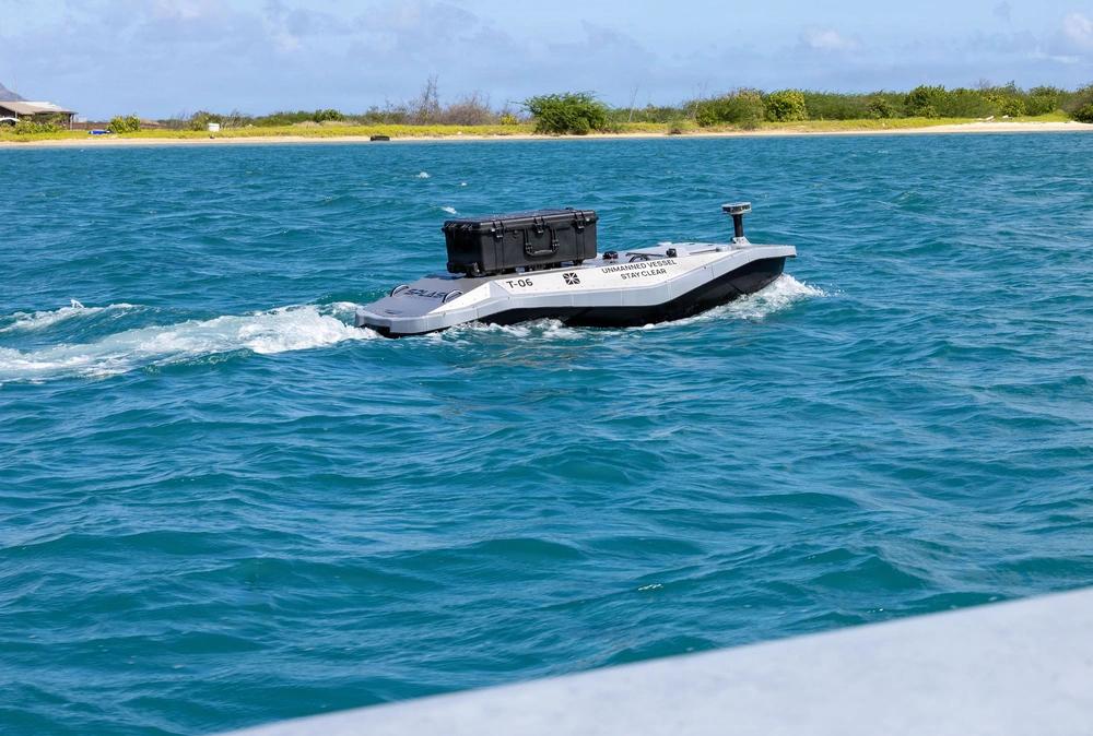  An unmanned surface vessel moves out to sea from Sand Island near Honolulu, Hawaii, to deliver parts for a 3D printer aboard the Wasp-class amphibious assault ship USS Essex (LHD-2) during Exercise Rim of the Pacific 2026, July 7, 2026. Although there is no human operator aboard such a vessel, there is a human footprint—and often a very large one—needed to operate and maintain an “unmanned system,” the author says. Credit: U.S. Navy photo by Mass Communications Specialist 3rd Class Charles Harbour
