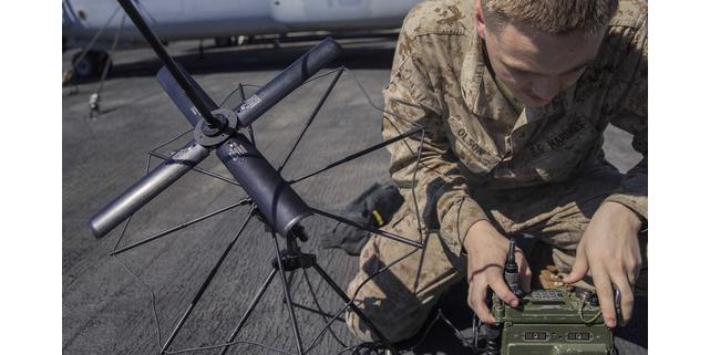 A U.S. Marine Corps Lance Corporal is assembling an AN/PRC-117G radio on the flight deck of an amphibious assault ship. The Mobile User Objective System (MUOS) adds updated firmware to the radio, giving warfighters more advanced satellite communications.” Credit: U.S. Marine Corps photo by Lance Cpl. Tawanya Norwood.